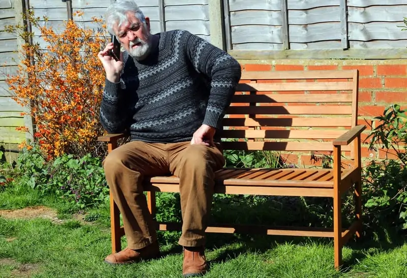 old man on a wooden bench holding a mobile phone listening to instructions